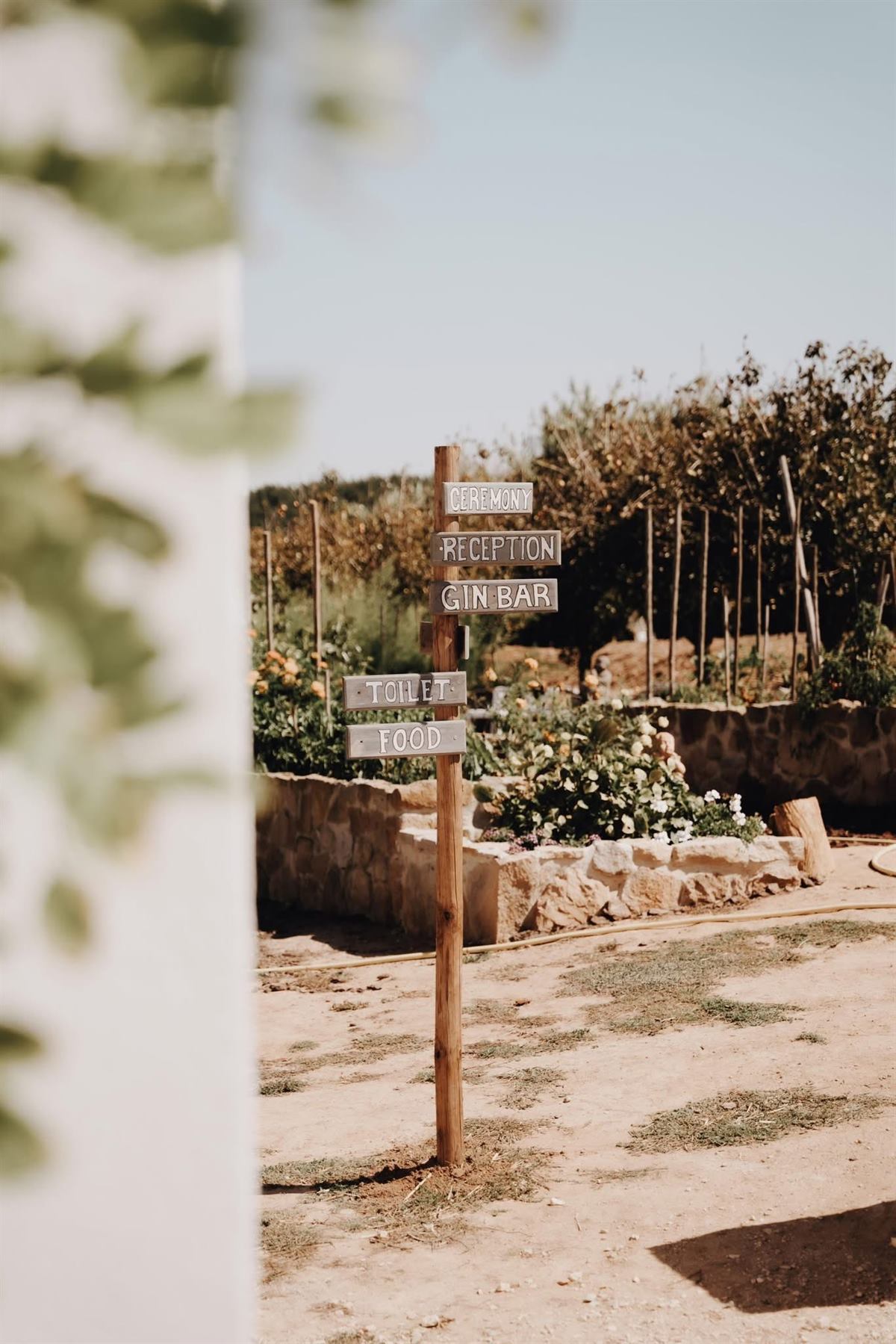 Rustic wooden directional signage for a wedding venue — Ceremony, Reception, Gin Bar, Toilet, Food — styled in a stone-walled garden setting