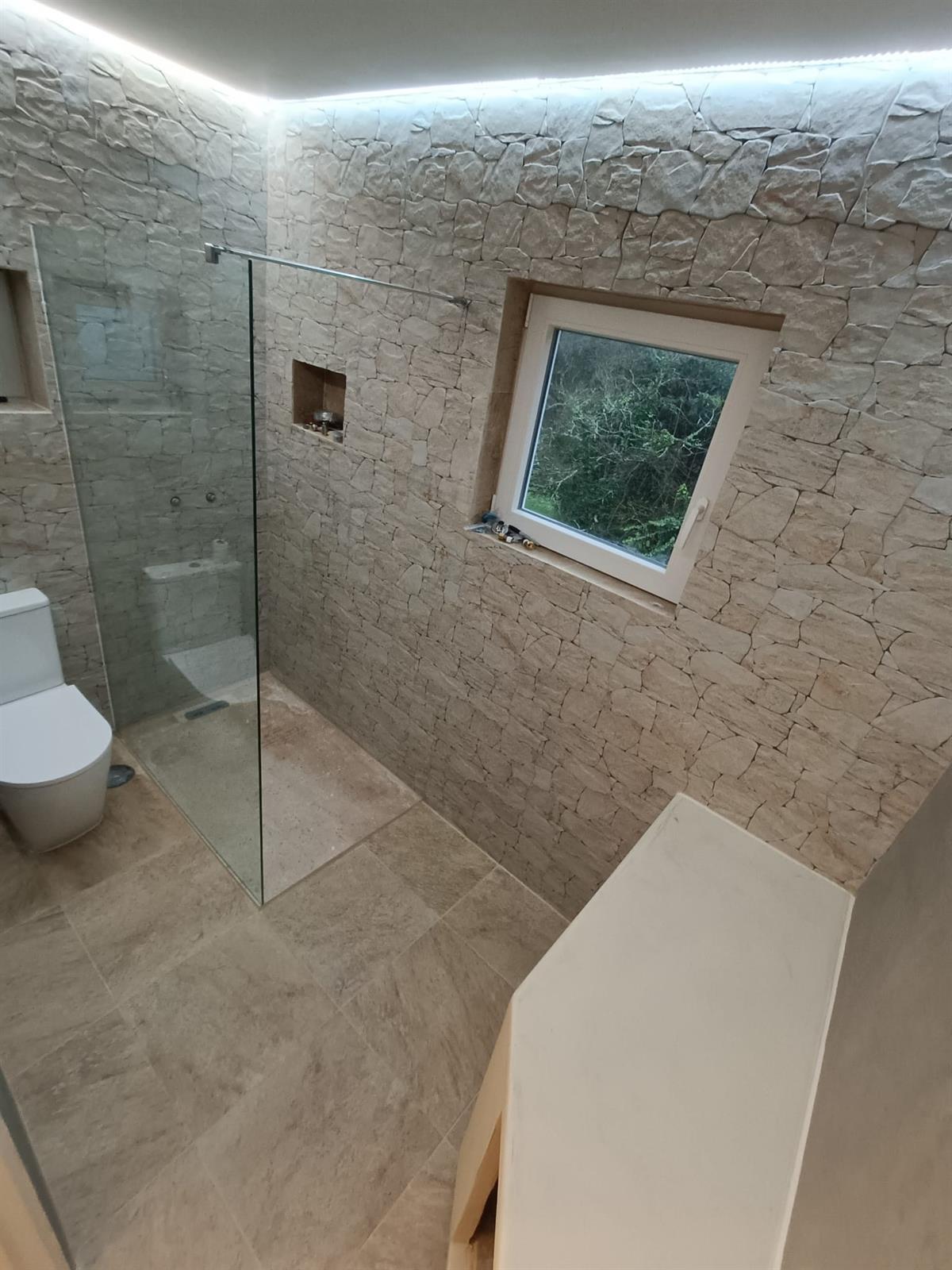 A finished stone-clad bathroom renovation on the Silver Coast &mdash; walk-in shower with glass screen, textured stone walls and a timber-framed window, completed by Justin Sterrenberg