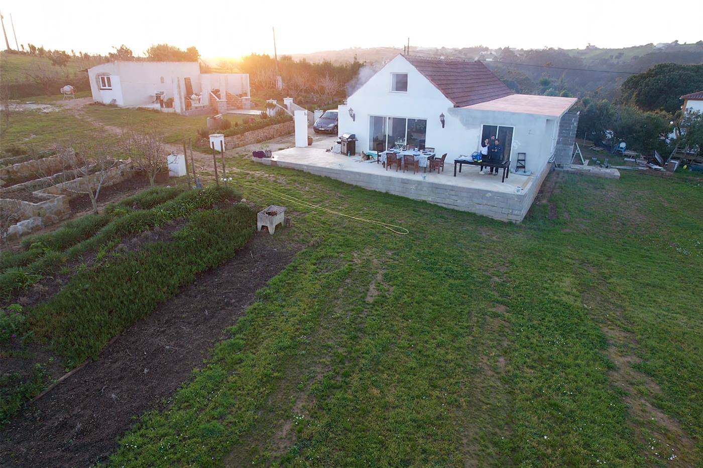 Aerial view of a finished white Silver Coast villa with a poured concrete terrace, outdoor dining and surrounding gardens at sunset